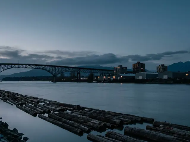 A wide river at dusk with a log boom along the bank, a steel truss bridge and low buildings with lit windows on the far shore.